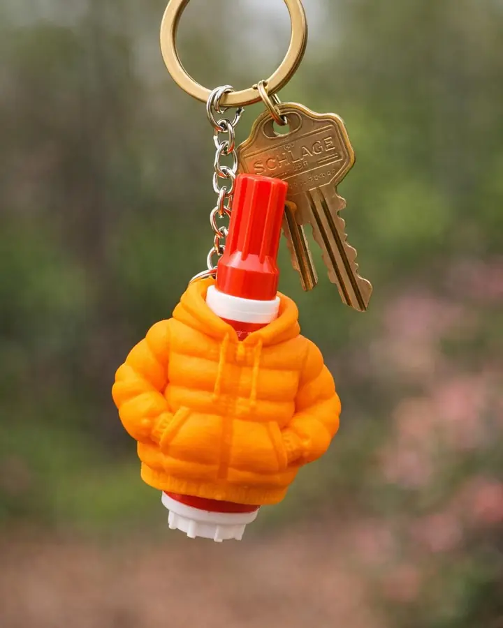 Orange puffer jacket charm holding a mini stain remover pen, on a keyring over warm wood