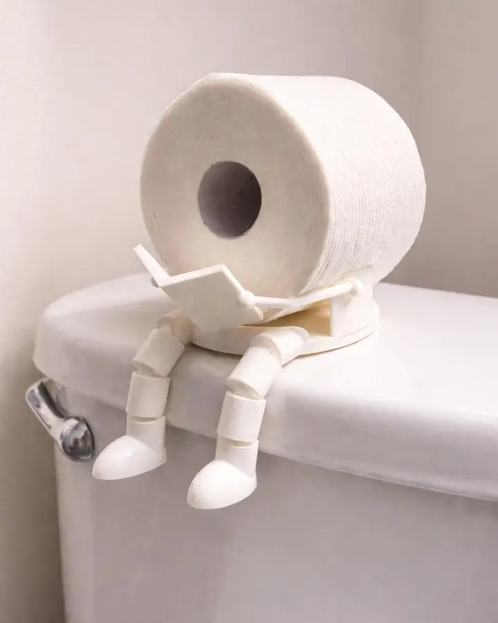White 3D printed figure with jointed legs sitting on a toilet tank, reading, holding a paper roll